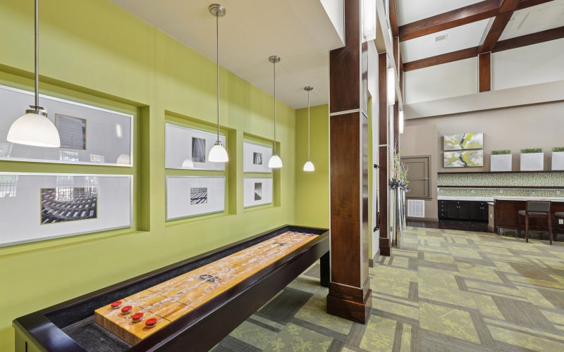 Clubhouse game room at Stone Pointe apartments featuring a wooden shuffleboard table, accent lighting, green walls, and high ceilings.